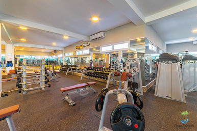 Bright modern indoor gym with mirrored walls, rows of dumbbells and benches, weight plates and resistance machines on rubber flooring — spacious fitness center workout area