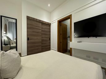 Cozy modern bedroom interior with white quilted bed, full-length mirror, wood sliding closet doors, open wooden doorway to an ensuite bathroom, and a wall-mounted flat-screen TV — neutral, minimalist decor.
