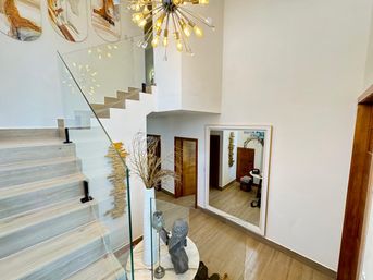 Bright modern entry foyer with light wood staircase and glass railing, sputnik chandelier overhead, large wall mirror, and decorative vases and sculptural accents on a round table.