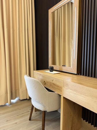 Cozy light-wood vanity with mirror, beige upholstered chair and floor-length tan curtains in a modern bedroom interior.