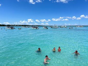 Sunny tropical sandbar with crystal-clear turquoise water, dozens of people wading and swimming around anchored dayboats and catamarans under a bright blue sky with puffy white clouds