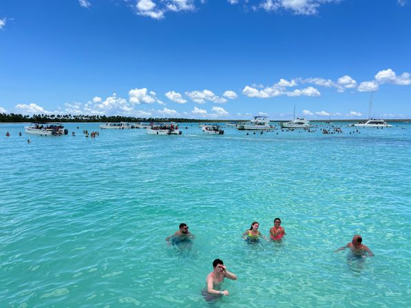 Sunny tropical sandbar with crystal-clear turquoise water, dozens of people wading and swimming around anchored dayboats and catamarans under a bright blue sky with puffy white clouds