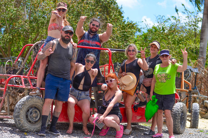 Group of eight adults in summer clothes and sunglasses posing and flashing peace signs beside a red off-road dune buggy with muddy tires under palm trees on a sunny tropical dune buggy tour excursion