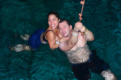 Smiling couple in swimwear holding a rope while floating in clear turquoise water, playfully enjoying a tropical swim