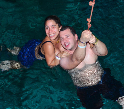 Smiling couple in swimwear holding a rope while floating in clear turquoise water, playfully enjoying a tropical swim