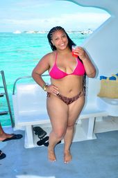 Smiling woman with braided hair in a pink bikini top and zebra-print bottoms holding a red drink on a boat deck over turquoise tropical water