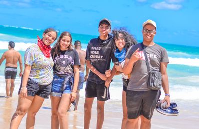 Five friends smiling and posing on a sunny tropical beach with turquoise waves and sandy shoreline, wearing casual summer clothes, bandanas and sandals for a playful beach day