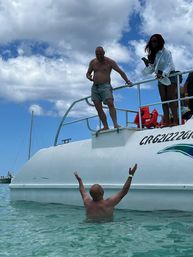 Two men enjoying a tropical boat excursion — one standing on a white boat platform reaching toward a friend in clear turquoise water as others watch from the deck under a bright blue sky with clouds.
