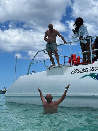 Two men enjoying a tropical boat excursion — one standing on a white boat platform reaching toward a friend in clear turquoise water as others watch from the deck under a bright blue sky with clouds.