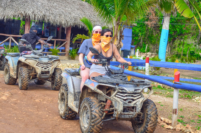 Muddy tropical ATV ride: two bandana‑clad riders on a quad passing palm trees and a thatched‑roof hut along a dirt trail.
