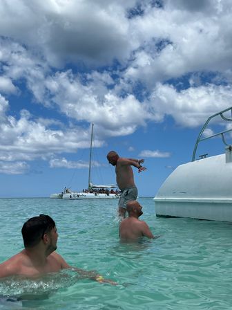 Three men playing in shallow turquoise water beside a white boat, one mid-jump off a platform with a sail catamaran and puffy clouds under a bright blue sky
