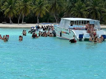 Groups of people socializing and swimming around anchored tour boats in a crystal-clear turquoise lagoon off a palm-fringed tropical beach