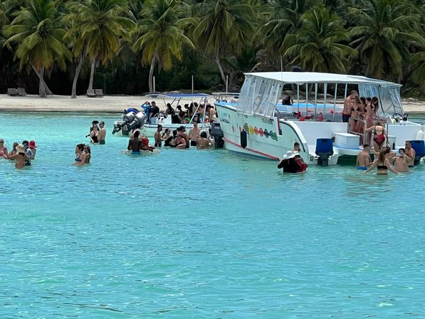 Groups of people socializing and swimming around anchored tour boats in a crystal-clear turquoise lagoon off a palm-fringed tropical beach
