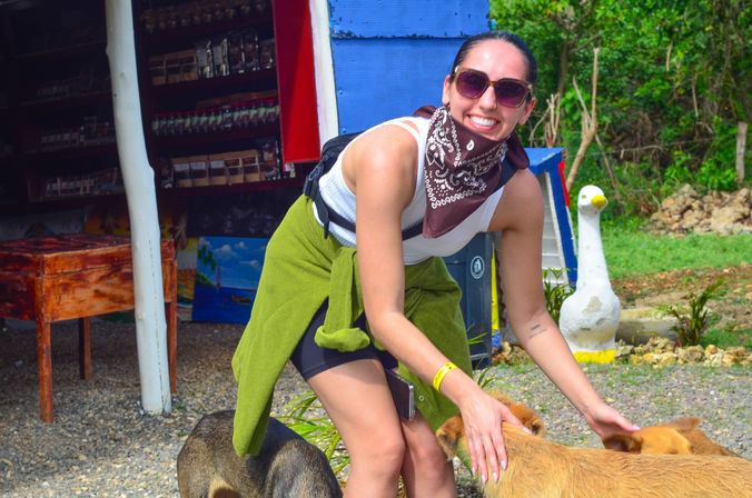 Smiling traveler in sunglasses and bandana petting friendly dogs outside a colorful roadside souvenir stand surrounded by tropical greenery.