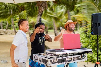 Three smiling men DJing and dancing at a tropical beach party under palm trees, red laptop on a DJ mixer case and a speaker nearby