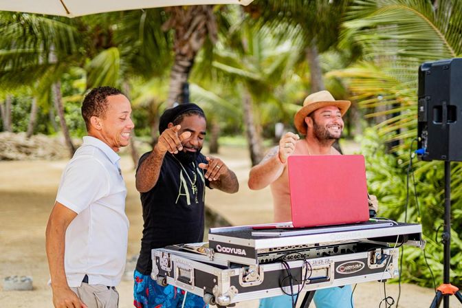Three smiling men DJing and dancing at a tropical beach party under palm trees, red laptop on a DJ mixer case and a speaker nearby