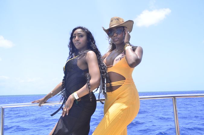 Two women posing on a boat railing with bright blue tropical ocean and sky behind them — one in a black mesh cover-up, the other in a yellow cutout dress, straw cowboy hat and sunglasses.