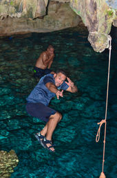 Person mid-air jumping from a rope swing into a turquoise underground limestone cave pool (cenote), with another swimmer wading in the background.