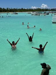 People wading and cheering in a shallow turquoise tropical lagoon with palm-lined shore and anchored tour boats under a bright blue sky