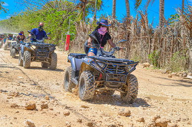 Off-road fun: group of ATV riders splashing through a muddy tropical dirt trail lined with palm trees under a bright blue sky