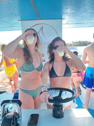 Two women in bikinis sipping yellow cocktails aboard a sunny party boat, one holding the helm, tropical coastline and other passengers visible in the background