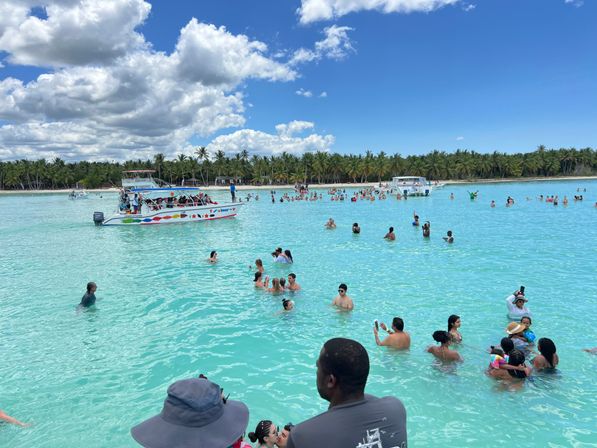 Crowded turquoise lagoon with tourists wading and taking photos, colorful tour boats nearby, palm‑lined tropical beach and blue sky with puffy clouds