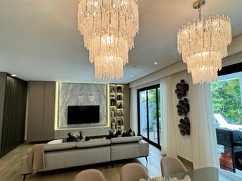 Sleek modern living room with cascading crystal chandeliers, marble TV wall, white leather sectional and sliding glass doors opening to a tropical patio