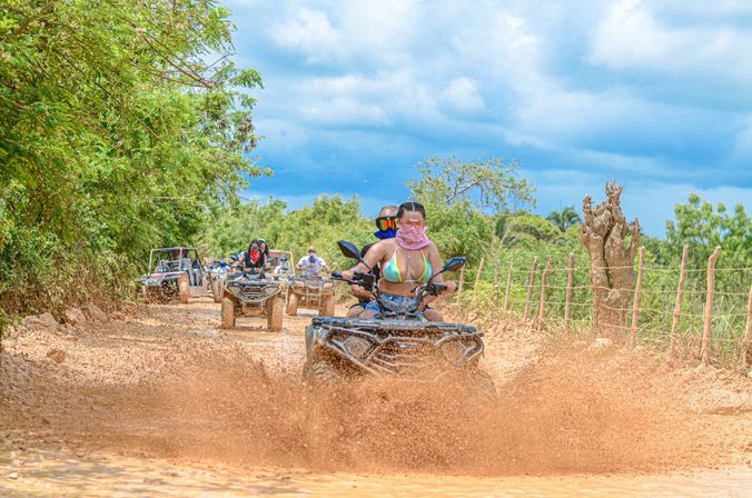 Group of ATV riders splashing through a muddy tropical dirt trail, lead rider in a colorful bikini top and face covering, lush green trees and a rustic fence under a dramatic blue sky.