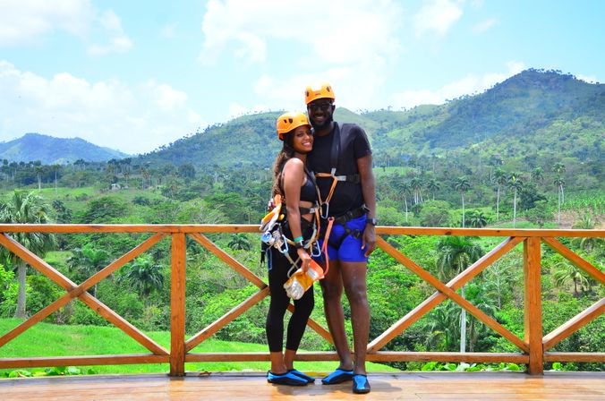Couple in helmets and zipline harnesses pose on a wooden platform, smiling against a lush tropical valley of palm trees and rolling green hills.