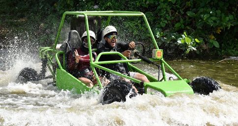 Two helmeted riders in a bright green off-road dune buggy splashing through whitewater in a tropical river, surrounded by lush green foliage on a joyful adventure tour.