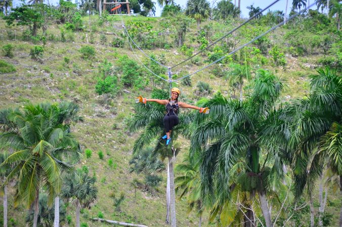 Person ziplining over a tropical hillside and palm tree canopy, wearing a helmet, harness and gloves with arms outstretched — outdoor adventure scene.