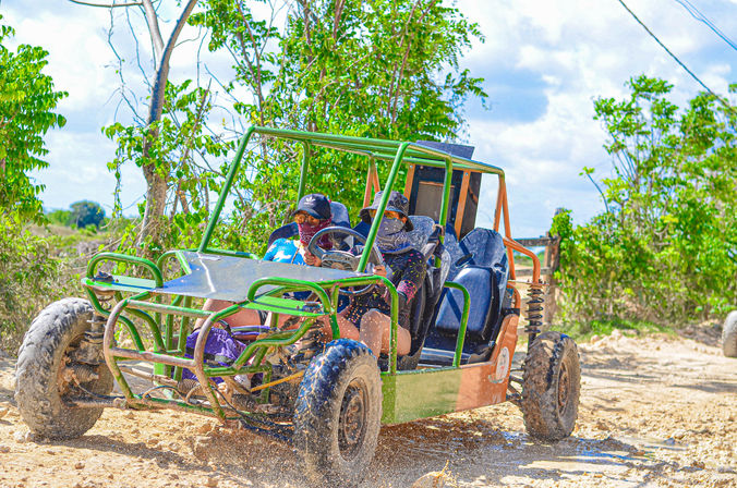 Two riders in a green off-road dune buggy kicking up mud and sand on a sunny tropical trail, surrounded by green trees — outdoor adventure ride.