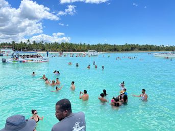 Shallow turquoise lagoon dotted with swimmers and colorful tour boats, palm‑fringed sandy beach and bright blue sky with fluffy clouds