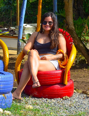 Smiling woman in sunglasses and a black tank top lounges cross-legged on a red-and-yellow painted tire chair in a sunny tropical outdoor setting with blue painted tires and green foliage