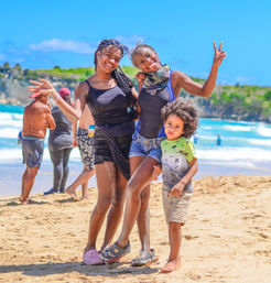 Two young women and a small child smiling and posing on a sunny sandy beach with turquoise waves and green coastal cliffs in the background, one woman flashing a peace sign.
