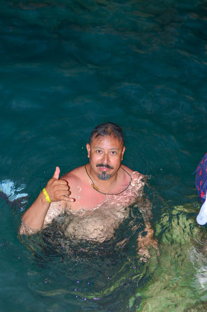 Smiling swimmer in deep teal water flashing a shaka (hang‑loose) gesture, wearing a yellow wristband and necklace.