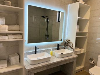 Spa-like modern bathroom interior with marble countertop, two white vessel sinks, matte-black faucets, illuminated backlit mirror, open shelving of folded white towels, and a walk-in shower with black rain showerhead.