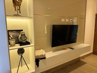 Modern apartment living room entertainment wall with wall-mounted flat-screen TV, beige tiled feature wall and floating media console; illuminated built-in shelves with warm LED lighting showcasing a cheeky monkey sculpture, tripod globe, wooden horse figurine, jar and a box of white roses, remote on the shelf.