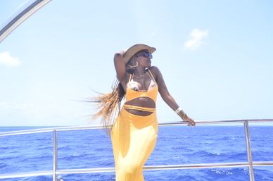 Person in a straw hat and sunglasses wearing a yellow cut-out dress, leaning on a yacht railing with long braids blowing over vivid blue ocean under a clear sky, tropical vacation vibe.