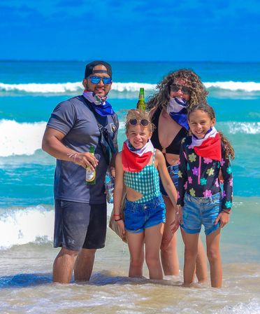 Family of four smiling in shallow surf on a sunny tropical beach with turquoise waves, colorful bandanas, sunglasses and casual swimwear — lively family beach day.