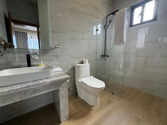 Modern marble bathroom interior with a vessel sink on a marble vanity, white toilet, glass walk-in shower with black fixtures, wood-look tile floor and small window.