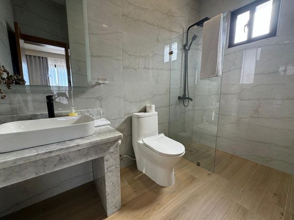 Modern marble bathroom interior with a vessel sink on a marble vanity, white toilet, glass walk-in shower with black fixtures, wood-look tile floor and small window.