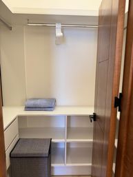 Minimalist walk-in closet with white built-in shelves and cubbies, chrome hanging rod holding stacked white hangers, folded gray towels on a shelf, gray fabric hamper and a wooden door