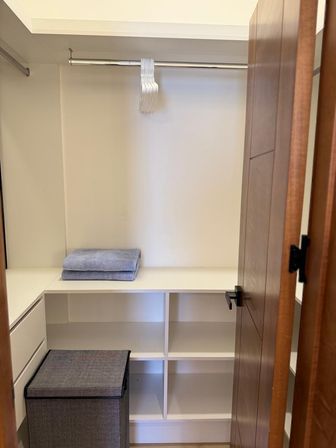 Minimalist walk-in closet with white built-in shelves and cubbies, chrome hanging rod holding stacked white hangers, folded gray towels on a shelf, gray fabric hamper and a wooden door