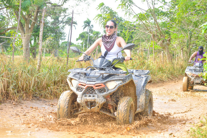 Smiling rider in sunglasses and bandana driving an ATV through a muddy tropical off-road trail, mud splashing with palm trees and another rider in the background.