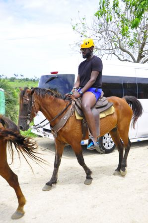 Rider in a yellow helmet and sunglasses on a brown horse, wearing a black shirt and blue shorts, trotting along a sandy trail with a white van parked nearby and tropical greenery and palm trees in the background.