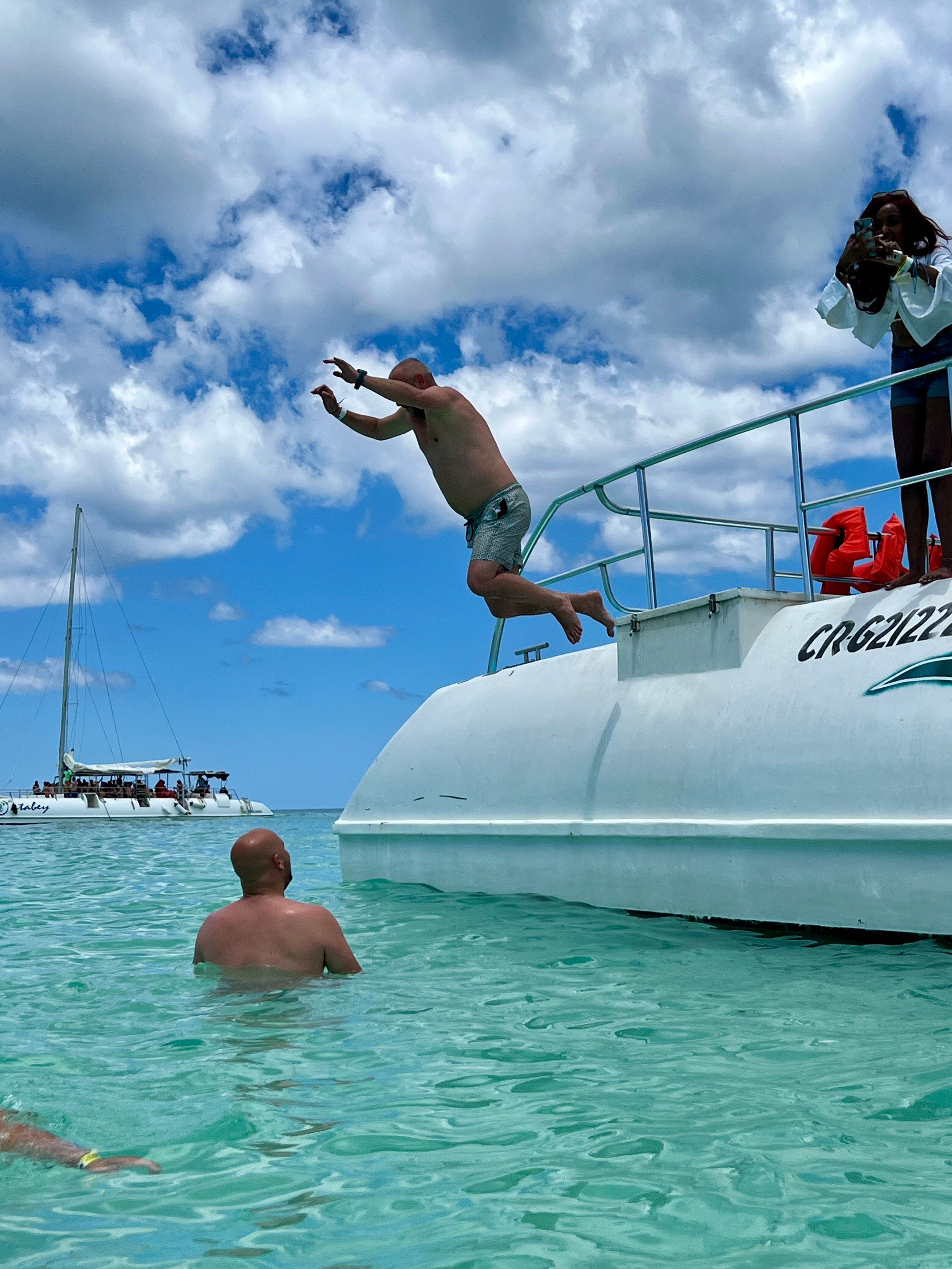 Man mid-air leaping off a white boat into crystal-clear turquoise tropical water, another swimmer watching in the shallow sea with a sailboat on the horizon under a bright partly cloudy sky.