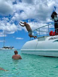 Man mid-air leaping off a white boat into crystal-clear turquoise tropical water, another swimmer watching in the shallow sea with a sailboat on the horizon under a bright partly cloudy sky.