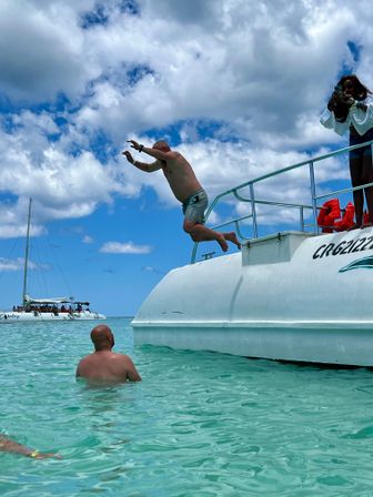 Man mid-air leaping off a white boat into crystal-clear turquoise tropical water, another swimmer watching in the shallow sea with a sailboat on the horizon under a bright partly cloudy sky.