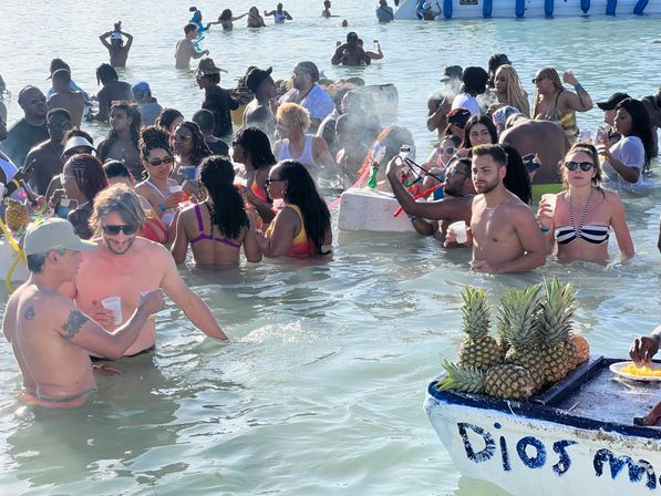 Festive sandbar beach party with a crowd in swimsuits mingling in shallow turquoise water around a floating bar/boat topped with pineapples, drinks, inflatables and a hookah on a sunny day.